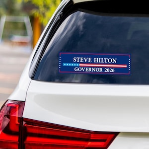 May include: A rectangular bumper sticker on a car window. The sticker has a dark blue background with white text that reads "STEVE HILTON GOVERNOR 2026". The text is framed by a red border and white stars.