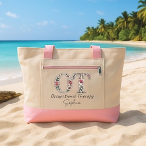 May include: A beige tote bag with pink accents, featuring the letters "OT" decorated with floral designs. The bag has a zippered pocket and handles, with the words "Occupational Therapy Sophia" printed below.