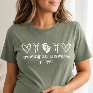 Puede incluir: Una camiseta de manga corta color verde oliva con un estampado gráfico blanco. El diseño incluye dos corazones, dos lazos y un par de huellas de bebé, con el texto "growing an answered prayer" impreso en una fuente blanca sin remates.