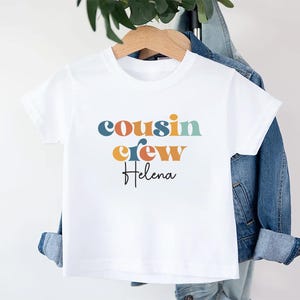 May include: White t-shirt with the text "cousin crew" in colorful letters and the name "Helena" written below in black.