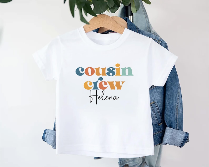 May include: White t-shirt with the text "cousin crew" in colorful letters and the name "Helena" written below in black.