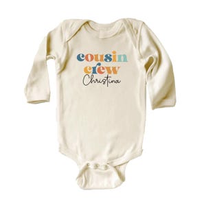 May include: A cream colored long-sleeved baby onesie with the text "cousin crew Christina" in colorful retro font.