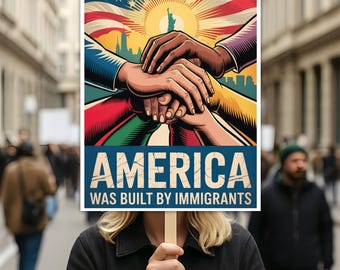 May include: A person holding a sign with the text "AMERICA WAS BUILT BY IMMIGRANTS". The sign features an illustration of clasped hands, the American flag, and the Statue of Liberty. The background shows a crowd of people.