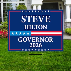 May include: A blue and red campaign sign on a metal stake, displayed on a green lawn. The sign features the name "STEVE HILTON" in white, with "GOVERNOR 2026" below. A blue and red stripe design is in the middle, with white stars.