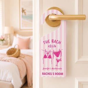 May include: A pink and white striped door hanger featuring the text THE BACH club, Jennifer's Bachelorette, and RACHEL'S ROOM. The design includes a graphic of a bikini and a cocktail glass hanging on a clothesline, printed in dark pink ink.