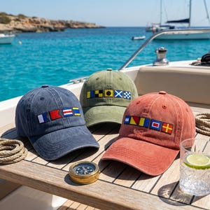 Peut inclure: Trois casquettes de baseball en bleu, vert olive et rouge terre cuite, chacune ornée d'une rangée de drapeaux de signalisation nautique brodés sur le panneau avant. Les casquettes ont une finition en tissu délavé sur un pont en bois.
