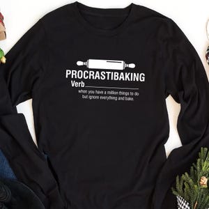 May include: Black long-sleeve t-shirt with white text that reads "PROCRASTIBAKING Verb. when you have a million things to do but ignore everything and bake." A white rolling pin is above the text.