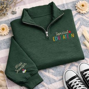May include: A dark green quarter-zip sweatshirt with the words "Special Education" embroidered in colorful letters. The sleeve features the embroidered text "Miss Jennifer" and a small apple and book design. Black and white sneakers are visible.