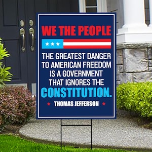 May include: A dark blue yard sign with the text "WE THE PEOPLE" in red, and a graphic of the American flag. The sign includes a quote by Thomas Jefferson about the Constitution.