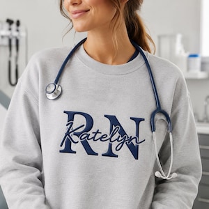 May include: A light gray crewneck sweatshirt featuring dark navy blue embroidery. The design displays the letters RN in a large block font with the name Katelyn written in a cursive script across the center. A dark navy blue stethoscope is draped around the neck.