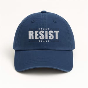 May include: A navy blue baseball cap featuring the word RESIST embroidered in white capital letters on the front. The text is centered between two horizontal lines, each flanked by five small white stars above and below the word.