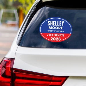 May include: An oval sticker on a car window displays the text "Shelley Moore West Virginia U.S. Senate 2026." The sticker is blue and red, with a white border. The car is white, with a red tail light visible.