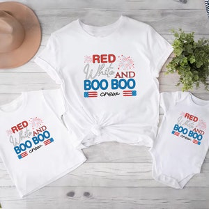 May include: Three white t-shirts with red, white, and blue text that reads "Red White and Boo Boo Crew". The shirts are on a white wooden surface.