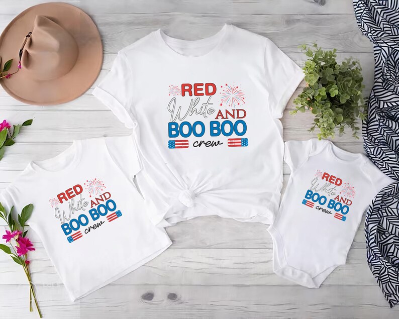 May include: Three white t-shirts with red, white, and blue text that reads "Red White and Boo Boo Crew". The shirts are on a white wooden surface.