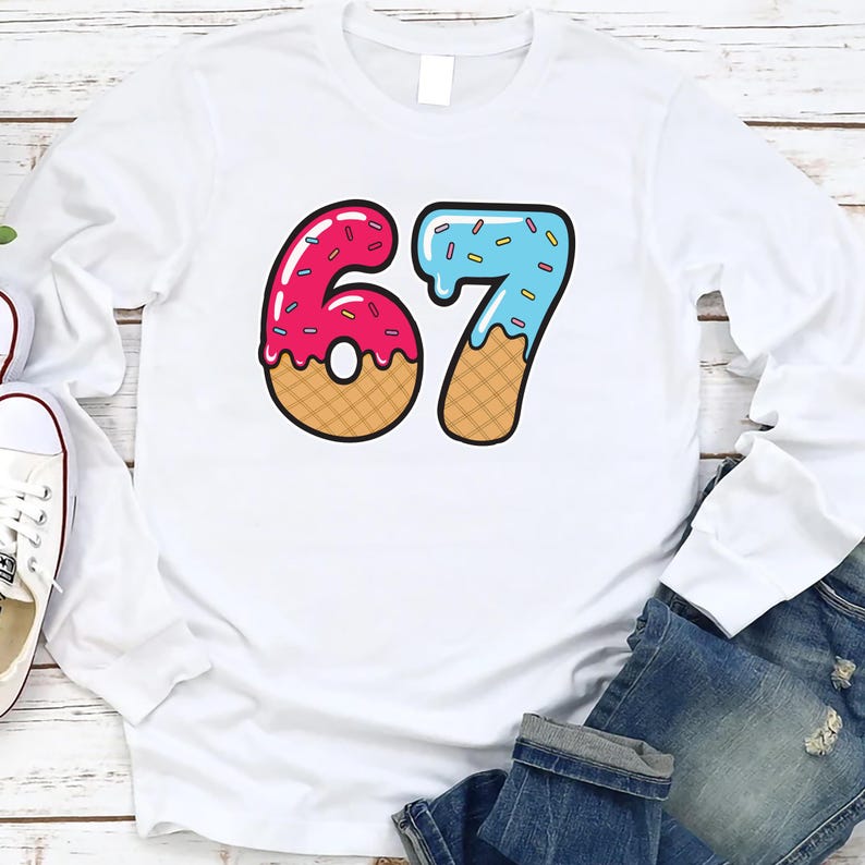 May include: White long-sleeve shirt featuring a colorful graphic of the number 67. The number is designed to look like a donut with pink frosting and sprinkles for the 6, and blue frosting and sprinkles for the 7.