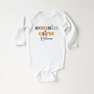 May include: White long-sleeve baby onesie with the text "cousin crew" in colorful retro-style letters and the name "Helena" written in cursive below.