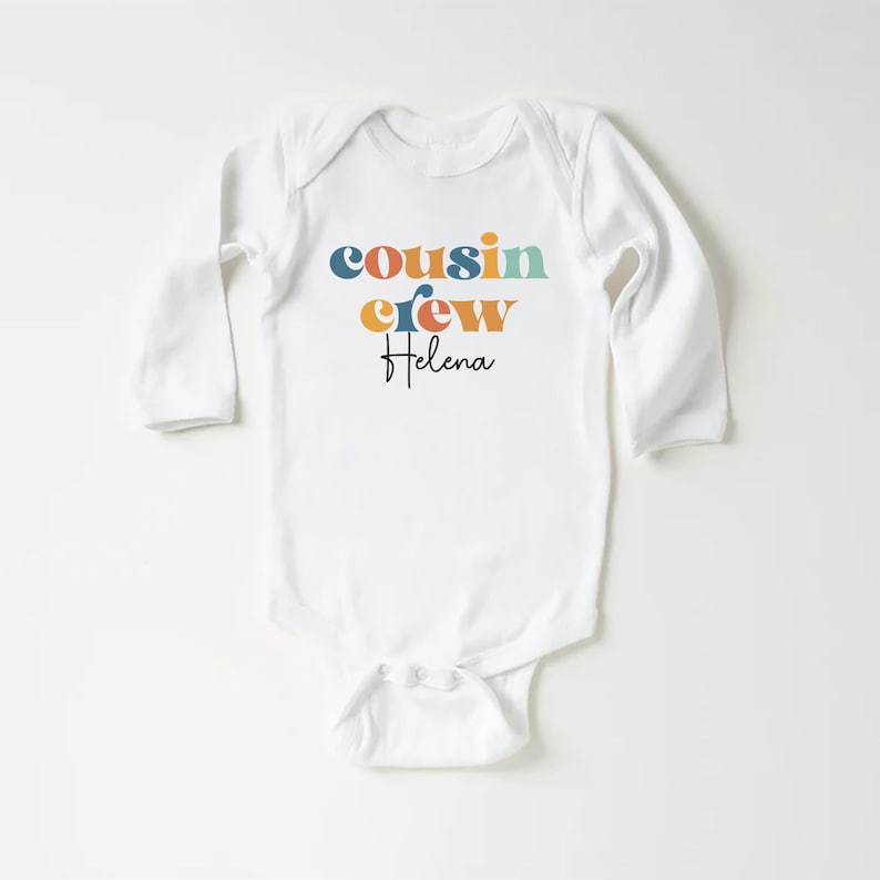May include: White long-sleeve baby onesie with the text "cousin crew" in colorful retro-style letters and the name "Helena" written in cursive below.