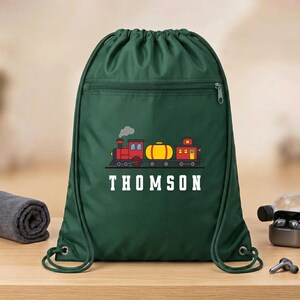 May include: A dark green drawstring backpack featuring a graphic of a red and yellow train with a smoke plume. Below the train, the name THOMSON is printed in white capital letters. The bag includes a horizontal zippered pocket across the front.