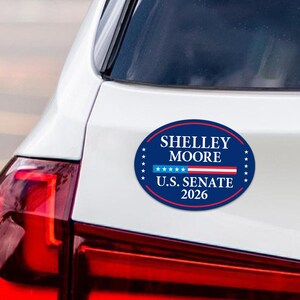 May include: An oval sticker on a car with the text "Shelley Moore U.S. Senate 2026". The sticker is dark blue with white text and a red and white stripe design. The car is white.
