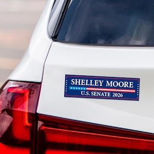 May include: A rectangular bumper sticker with the text "SHELLEY MOORE U.S. SENATE 2026" in white letters on a blue background with white stars and red and white stripes. The sticker is affixed to a white car.