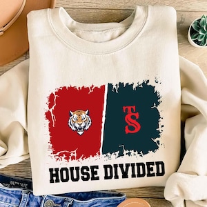 May include: Cream-colored sweatshirt with a graphic design. The design features a red and navy blue rectangle with a tiger and the letters "FS". The words "HOUSE DIVIDED" are printed in bold black letters below the design.
