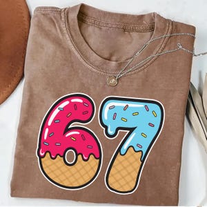 May include: A brown t-shirt with a large graphic of the number 67. The number 6 is pink with sprinkles and a waffle cone base. The number 7 is blue with sprinkles and a waffle cone base. A silver necklace is draped across the shirt.