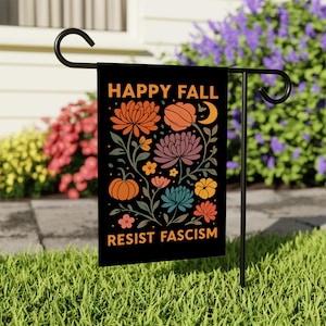 May include: Black garden flag with the words "HAPPY FALL" and "RESIST FASCISM" in orange text. The flag features a floral design with orange, purple, and blue flowers, pumpkins, and a crescent moon. The flag is displayed on a black metal stand in a garden.