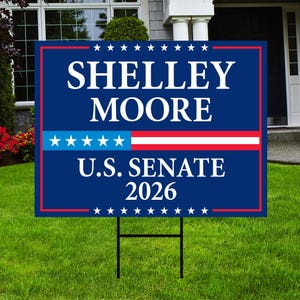 May include: A blue yard sign with white text reading "SHELLEY MOORE U.S. SENATE 2026". The sign features a red border and a stars and stripes design. It's set on a green lawn, supported by a metal stake.
