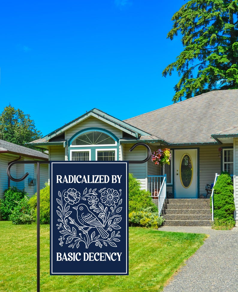 Puede incluir: Una bandera de jard&iacute;n azul marino con texto blanco y un dise&ntilde;o de p&aacute;jaro y flores. La bandera dice "RADICALIZED BY BASIC DECENCY". La bandera se muestra en un jard&iacute;n delantero con una casa y cielo azul en el fondo.