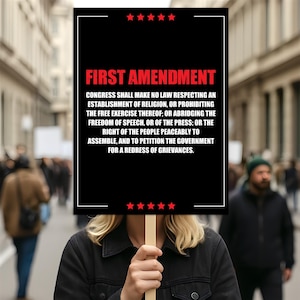 May include: A black sign with the words "FIRST AMENDMENT" in red, held by a person. The sign displays the text of the First Amendment, with red stars at the top and bottom. The background is a blurred street scene.