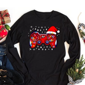 May include: Black long-sleeve shirt with a red and white video game controller design. The controller is decorated with Christmas lights and a Santa hat.