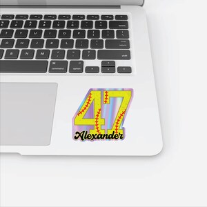 May include: A baseball-themed sticker with the number "47" in yellow, featuring red stitching details. The name "Alexander" is written below in a black script font. The sticker has a holographic background.