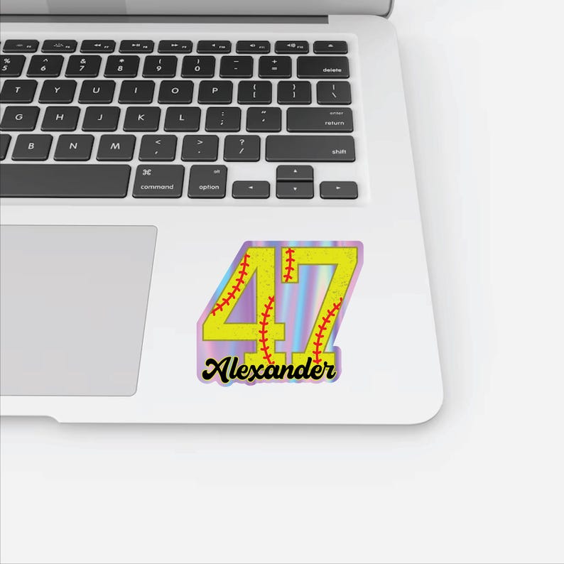 May include: A baseball-themed sticker with the number "47" in yellow, featuring red stitching details. The name "Alexander" is written below in a black script font. The sticker has a holographic background.