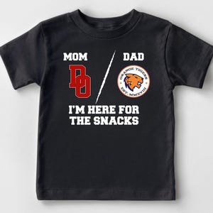 May include: Black t-shirt with white text that reads "Mom/Dad I'm here for the snacks". The shirt has a red "D" on the left side and a circular orange and blue logo with a tiger on the right side.