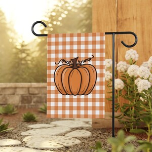 May include: A garden flag featuring a large orange pumpkin with the words "fuck trump" in a cursive font. The flag has a checkered orange and white background and is displayed on a black metal stand.