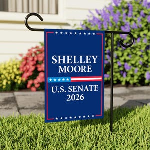 May include: A blue garden flag with the text "SHELLEY MOORE U.S. SENATE 2026" in white. The flag has a red border and a design of stars and stripes. It is displayed on a black metal stand in a grassy garden.