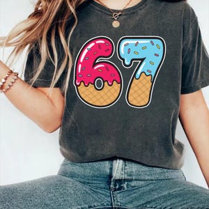 May include: Dark gray t-shirt featuring a colorful graphic of the number "67" designed to look like donuts. The "6" is pink with sprinkles, and the "7" is blue with sprinkles. The numbers have a waffle cone base.