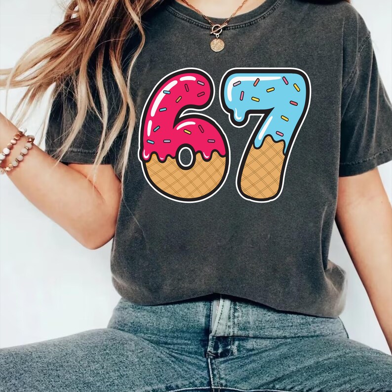 May include: Dark gray t-shirt featuring a colorful graphic of the number "67" designed to look like donuts. The "6" is pink with sprinkles, and the "7" is blue with sprinkles. The numbers have a waffle cone base.