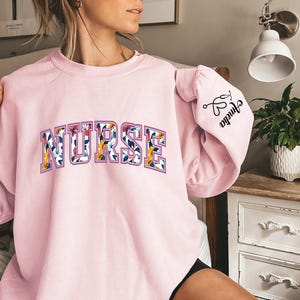 May include: Pink sweatshirt with the word "NURSE" in floral letters. The left sleeve has the text "Shrinka" with a pair of scissors and a heart.