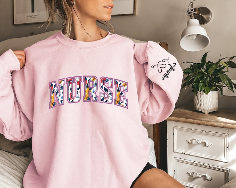 May include: Pink sweatshirt with the word "NURSE" in floral letters. The left sleeve has the text "Shrinka" with a pair of scissors and a heart.