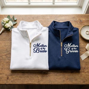 May include: A white quarter-zip sweatshirt with navy blue text reading Mother of the Bride and a navy blue quarter-zip sweatshirt with white text reading Mother of the Groom. Both garments feature gold-toned metal zippers and stand-up collars.