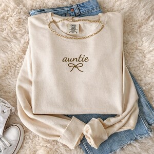 May include: Cream-colored sweatshirt with the word "auntie" and a bow graphic in brown. A gold chain necklace is draped around the neckline. The sweatshirt is laid on top of blue jeans and a fluffy white surface.