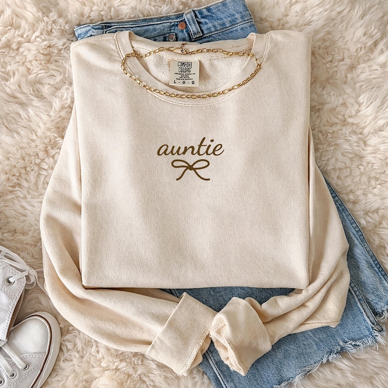 May include: Cream-colored sweatshirt with the word "auntie" and a bow graphic in brown. A gold chain necklace is draped around the neckline. The sweatshirt is laid on top of blue jeans and a fluffy white surface.