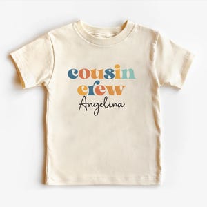 May include: A cream colored short-sleeved t-shirt with the text "cousin crew" in colorful retro font and the name "Angelina" in black script.