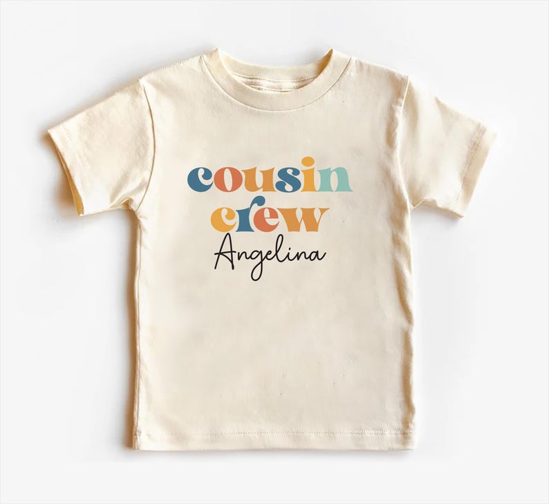 May include: A cream colored short-sleeved t-shirt with the text "cousin crew" in colorful retro font and the name "Angelina" in black script.