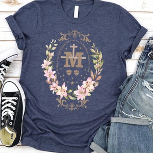 Op de afbeelding: Een marineblauw t-shirt met een bloemenkransontwerp met een gouden "M" met een kruis en twee harten. De tekst rond de krans luidt "O Maria, ontvangen zonder zonde, bid voor ons die onze toevlucht tot u nemen."