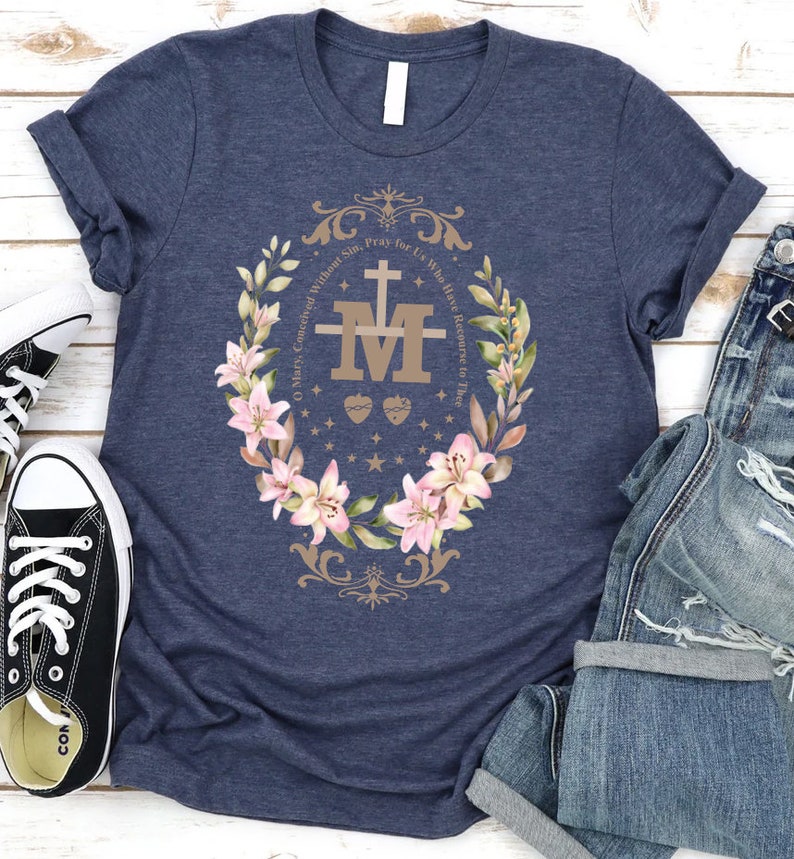 Op de afbeelding: Een marineblauw t-shirt met een bloemenkransontwerp met een gouden "M" met een kruis en twee harten. De tekst rond de krans luidt "O Maria, ontvangen zonder zonde, bid voor ons die onze toevlucht tot u nemen."