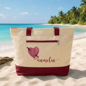 May include: Cream-colored tote bag with a burgundy base, handles, and zipper pocket. The bag features a heart-shaped yarn design with a knitting needle and the name "Tramelia" in a script font. The bag is on a sandy beach with a turquoise ocean in the background.