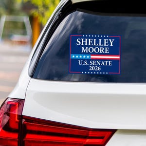 May include: A rectangular sticker on a car window displays the text "SHELLEY MOORE" in white capital letters above "U.S. SENATE 2026" in white. The text is set against a dark blue background with red and white stripes.