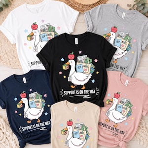 May include: A collection of five short-sleeve t-shirts in white, gray, black, navy, and light pink. Each shirt features a graphic of a white goose carrying school supplies, an apple on its head, and the text "SUPPORT IS ON THE WAY".